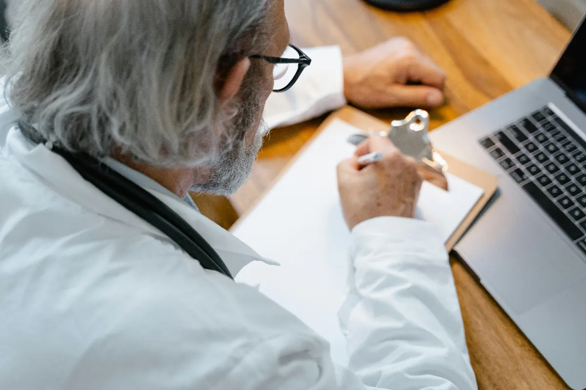 An elderly doctor in a white coat writing notes on a clipboard at a desk with a laptop. An elderly doctor in a white coat writing notes on a clipboard at a desk with a laptop.