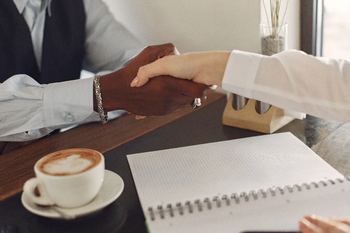 Two professionals shaking hands at a desk with a coffee cup and notebook, symbolizing business agreement. Two professionals shaking hands at a desk with a coffee cup and notebook, symbolizing business agreement.