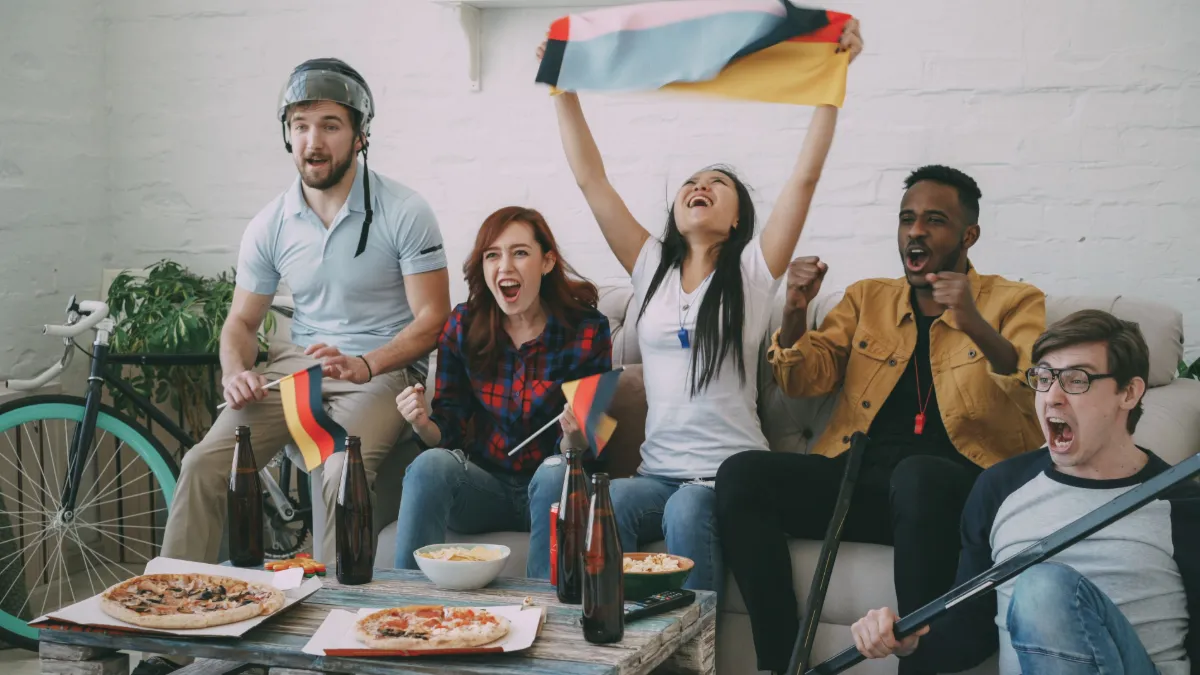 A diverse group of friends excitedly watches and cheers for a sports event indoors, holding flags.