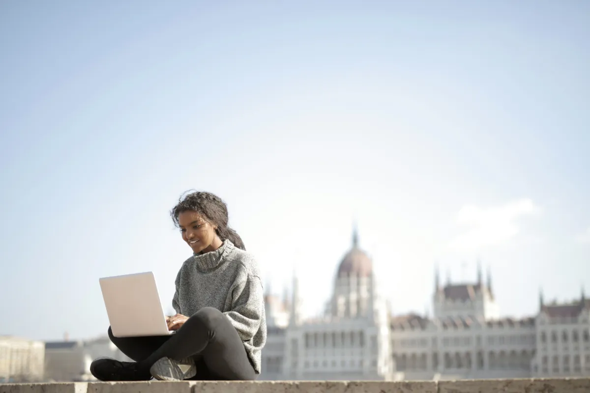 Young woman sitting outdoors with a laptop, working remotely against a blurred city landmark background on a sunny day.
