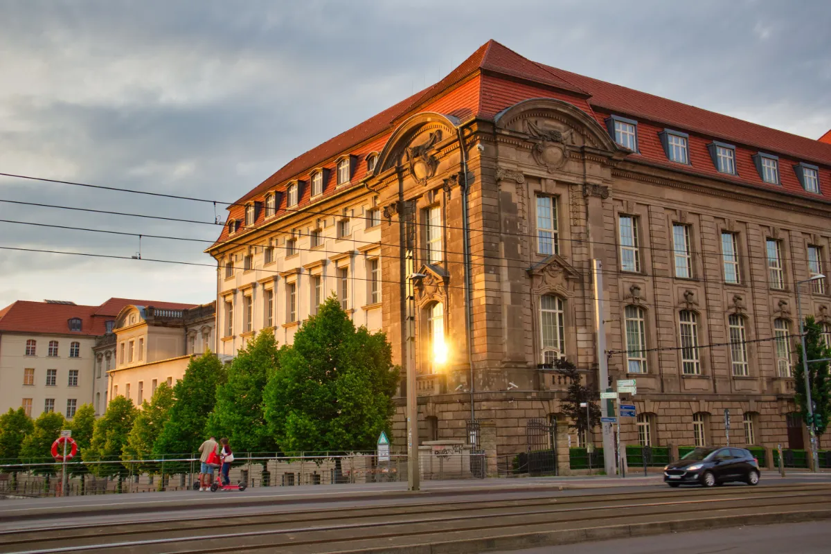 Historic European building with a red roof illuminated by golden sunset light, overlooking tram tracks and a city street.