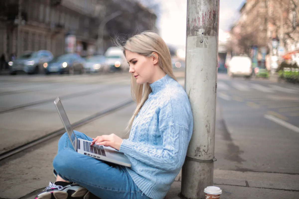Young woman sitting on a city sidewalk with a laptop, working remotely near tram tracks and blurred urban traffic.